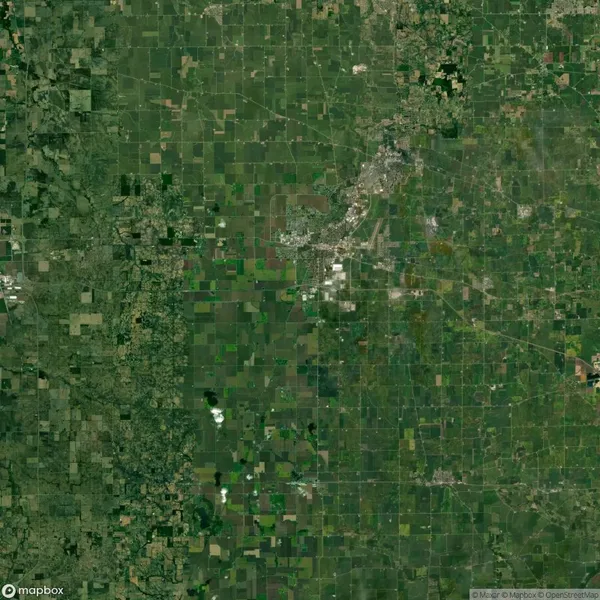 Aerial view of a large, patchwork landscape with green agricultural fields and scattered urban areas, featuring a grid-like pattern of roads and varying shades of green.