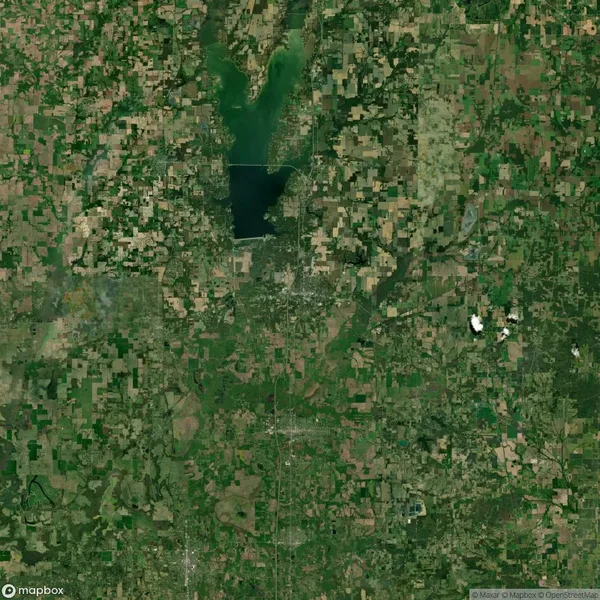 Aerial view of a patchwork landscape featuring a mix of green and brown fields. A large lake is visible at the top center with a grid-like pattern of roads and vegetation surrounding it.