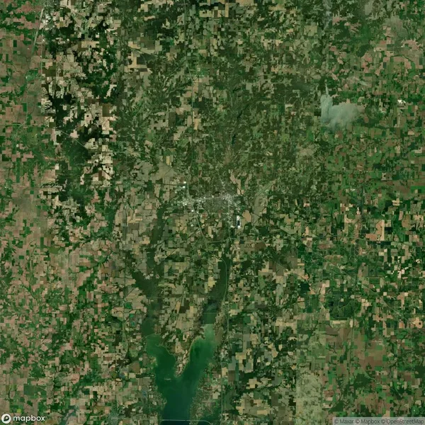 Satellite image showing a densely wooded and agricultural area surrounding a small town. A river runs from the lower section, widening into a body of water at the bottom of the image.