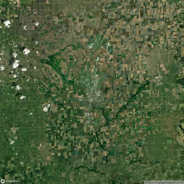 Aerial view of a patchwork landscape with fields and a central town, featuring green areas, roads, and scattered clouds.