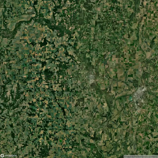 Aerial view of agricultural land with patchwork fields, forests, and a winding river cutting across the landscape. A town is visible on the right side of the image.