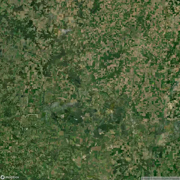 Aerial view of a large patchwork of agricultural fields, forests, and scattered small settlements. The landscape appears predominantly green and brown with varying field shapes and sizes.