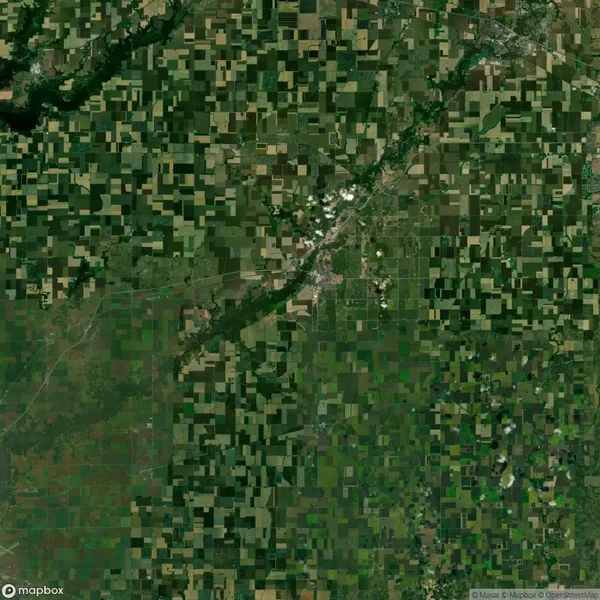 Aerial view of a rural area with a patchwork of green and brown agricultural fields. A small town is visible in the center, intersected by roads and surrounded by farmland.
