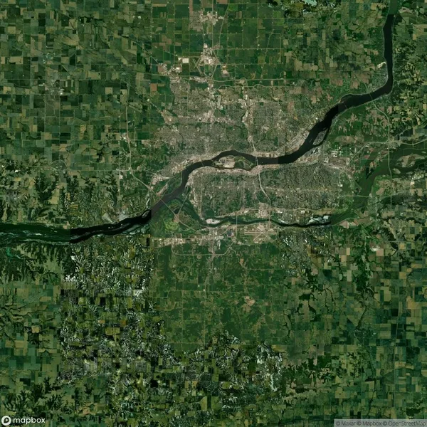Satellite image of a sprawling urban area divided by a river, surrounded by a mix of agricultural fields and forested areas.