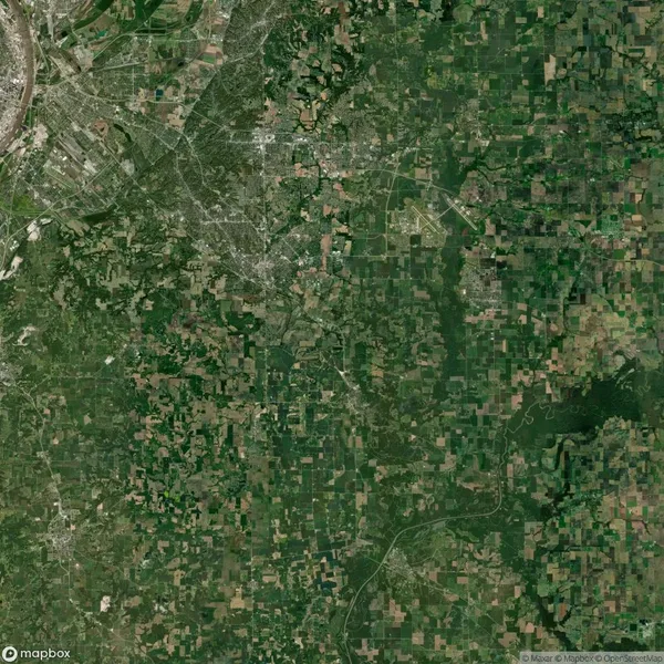 Satellite view of a landscape with a mix of urban and rural areas, showing a dense city on the top left and a patchwork of green fields and forests extending to the right and bottom.