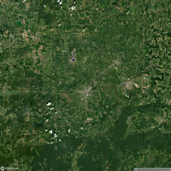Aerial view of a patchwork landscape with fields, forests, and scattered clouds. A town and roads are visible in the center.