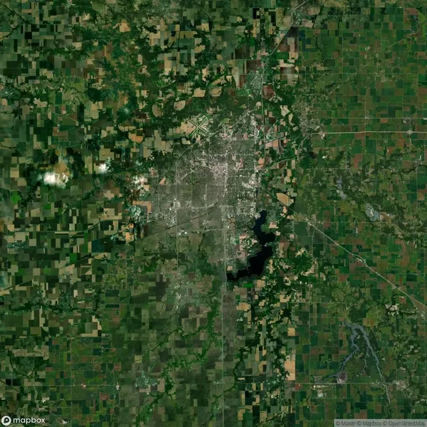 Satellite image of a landscape with a city center surrounded by farmland, roads, and a visible lake to the south. Sparse clouds are scattered across the area.