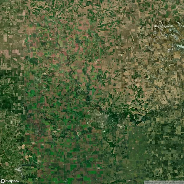 Satellite view of a large area showing a patchwork pattern of agricultural fields, with varying shades of green and brown, intersected by roads and a few scattered patches of wooded areas.