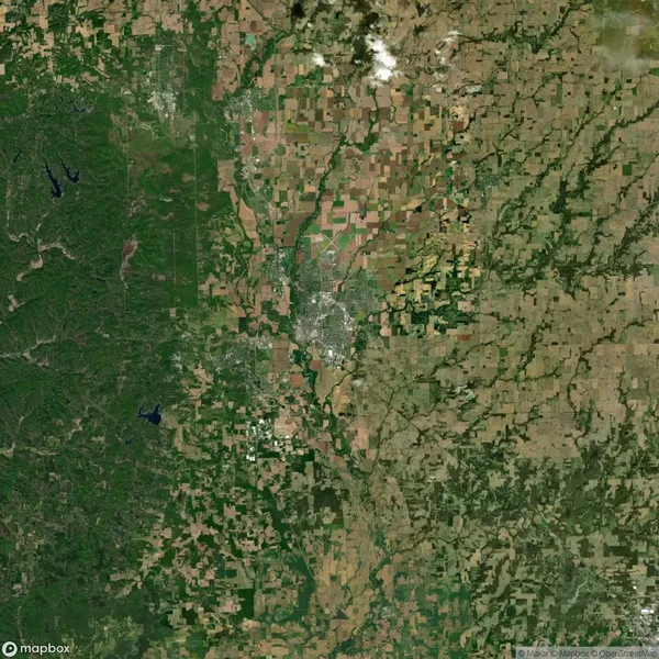 Satellite image showing a mix of farmlands, forests, and urban development. Central area is densely populated, surrounded by agricultural fields and patches of green forest.