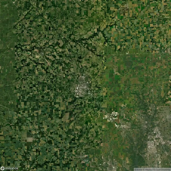 Aerial view of rural landscape with patchwork of green fields and small urban center in the middle. Roads and vegetation patterns are visible, dividing the landscape.