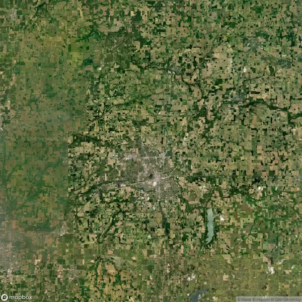 Satellite view of a landscape with a central urban area surrounded by a patchwork of agricultural fields and natural green spaces. Roads intersect the area in a grid-like pattern.