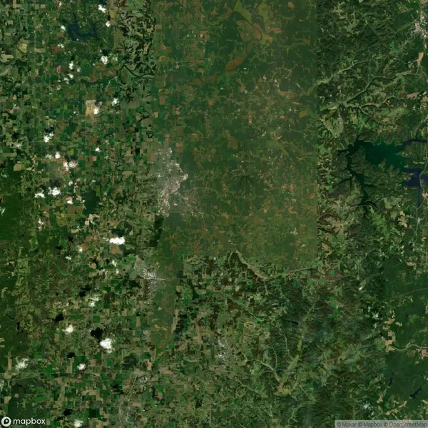 Satellite view of a diverse landscape with scattered clouds. Shows a mix of green fields, forests, and a central urban area.