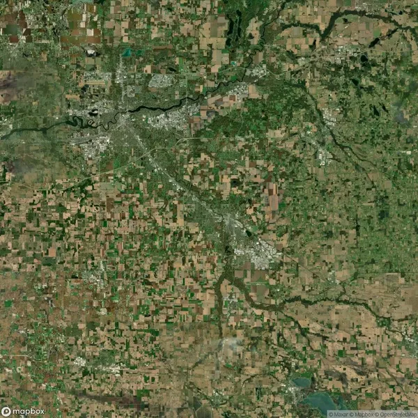 Satellite image showing a sprawling urban area surrounded by patches of green fields and agricultural land, intersected by rivers and small roads, under clear skies.