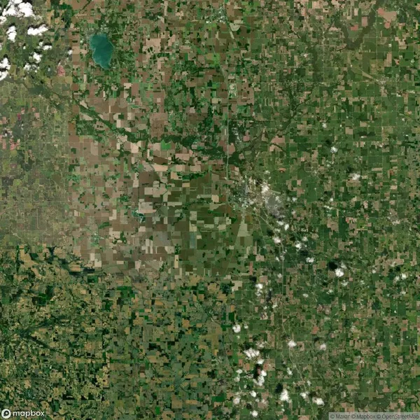 Aerial view of a large patchwork landscape with various shades of green and brown fields; a small blue body of water is visible in the top left corner.