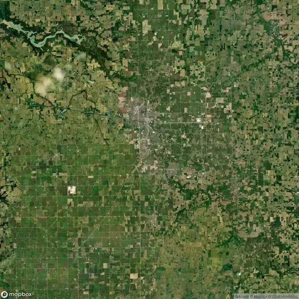 Aerial view of a rural landscape with a patchwork of green and brown fields, a river in the top left, and a densely built-up area in the center.