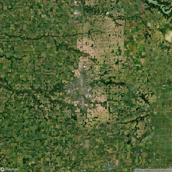 Aerial view of a vast green and brown patchwork landscape, likely farmland, with a dense urban area at the center. A river meanders on the right side.