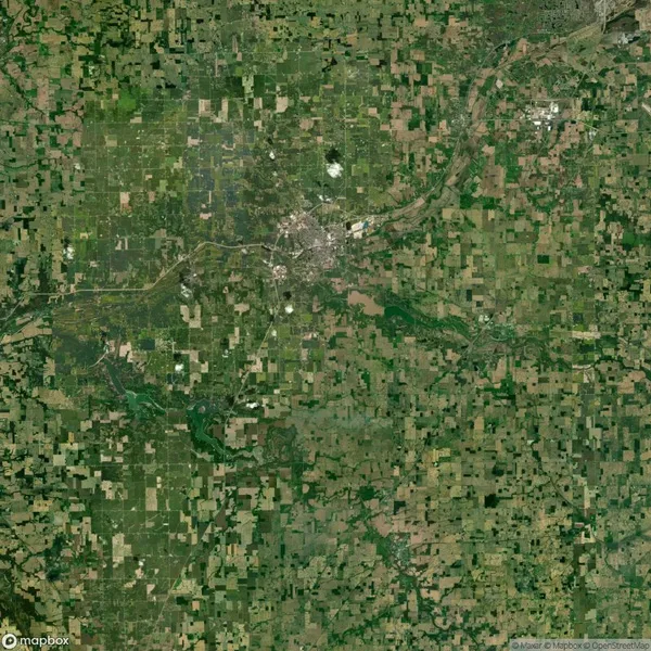 Satellite view of a rural landscape with a patchwork of green and brown fields, a central urban area with a river running through it, and a network of connecting roads.