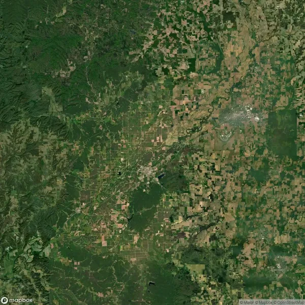 Aerial view of a landscape with mixed geography featuring green forests, agricultural fields, and a river winding through the terrain. A town or city is visible in the top-right area.