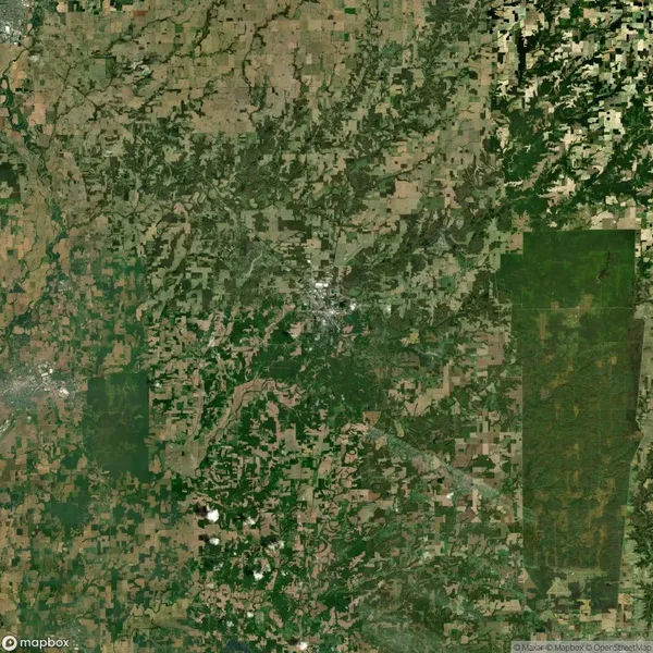 Aerial view of a patchwork landscape with varying shades of green and brown fields, intersected by roads, and denser vegetation in some areas. Sparse urban development is visible.