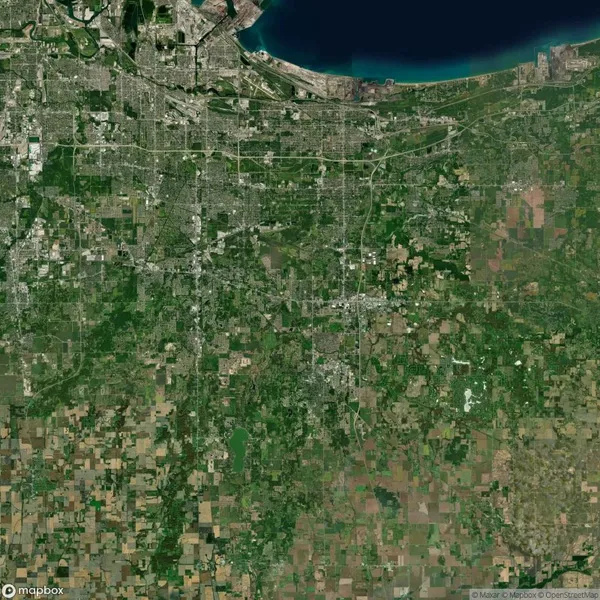 Satellite view of a landscape with a dense city grid near a body of water at the top and sprawling green fields extending towards the bottom.