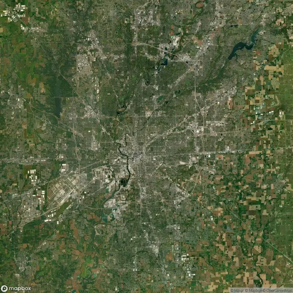 A satellite view of a large cityscape surrounded by green fields and patches of forest. A river runs through the city center, with various road networks and buildings visible throughout.