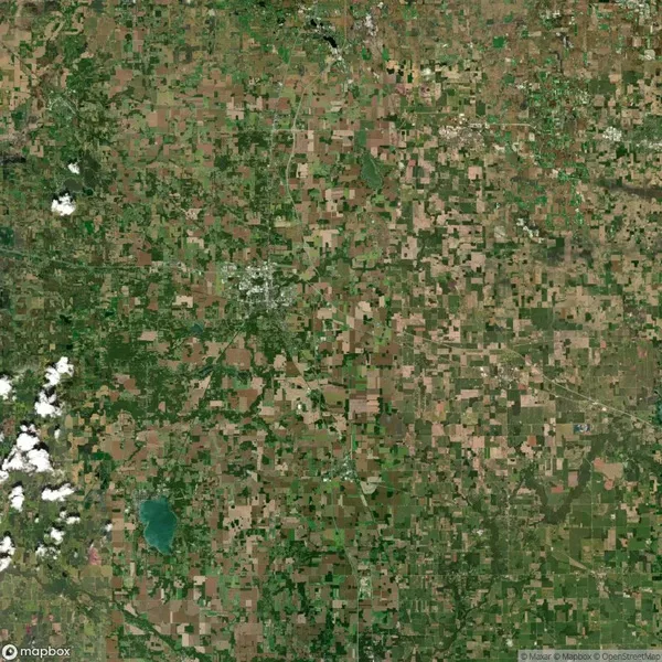 Aerial view of a rural landscape with a patchwork of agricultural fields, green areas, and scattered clouds in the bottom left corner.