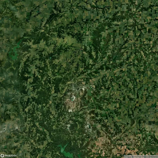 Aerial view of a landscape with dense green patches of forest and sprawling agricultural fields, divided by roads and natural features.