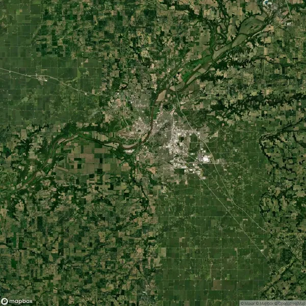 Aerial view of a city surrounded by rural farmland and a river. The city is located at the center with visible roads, while fields and natural landscapes extend outward.