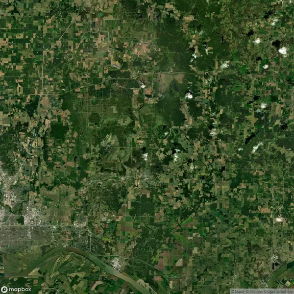 Satellite view of a landscape featuring a patchwork of agricultural fields, greenery, and scattered wooded areas. A river is visible in the lower part of the image.
