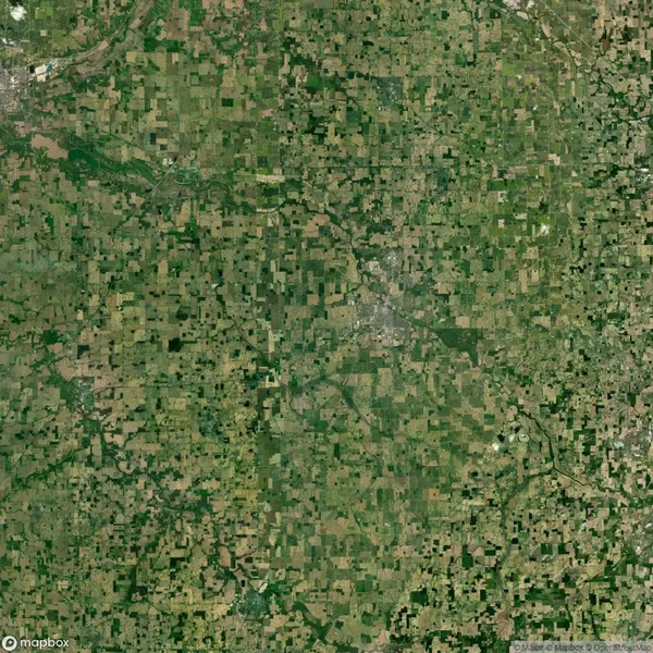 Aerial view of a vast, patchwork landscape with various shades of green and brown, indicating farmland and vegetation. Roads crisscross through the fields, and a small urban area is visible.