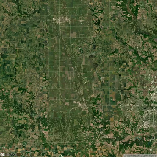 Aerial view of a rural landscape with a patchwork of agricultural fields, greenery, and scattered towns. A river runs through the center, and a grid of roads is visible throughout.