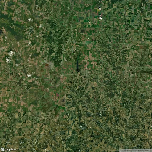 Aerial view of a rural landscape with green agricultural fields, small patches of forest, and a river meandering through the area. Several scattered clouds are visible in the sky.