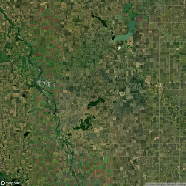 Aerial view of a patchwork landscape with varied shades of green and brown, indicating fields, forests, and water bodies. A river runs through the left side of the image.