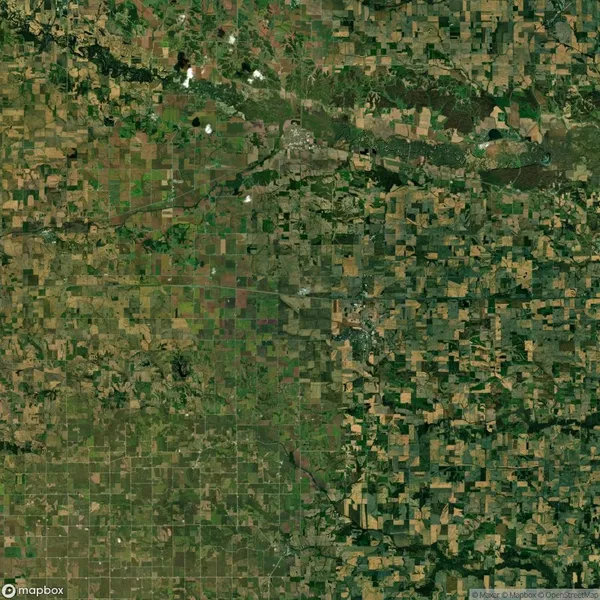 Aerial view of a patchwork landscape showing a grid of agricultural fields with varying shades of green and brown. A river or stream runs diagonally across the upper portion of the image.