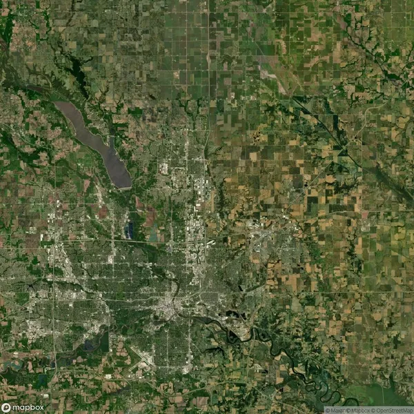 Satellite view of a landscape featuring a city at the center, surrounded by a mix of agricultural fields, patches of forest, and a river flowing at the bottom of the image.