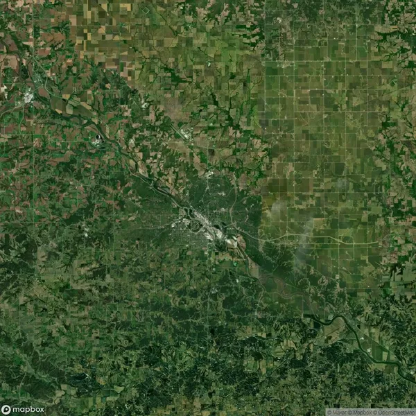 Satellite view of a green and brown landscape with patchwork fields and a winding river crossing horizontally. A densely built area is visible near the center.