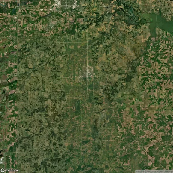 Aerial view of a vast landscape with patchwork fields and scattered patches of greenery. A winding river is visible at the top right, surrounded by a mix of rural and semi-urban areas.