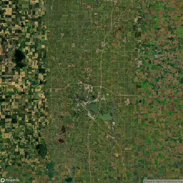 Aerial view of a town surrounded by a grid of agricultural fields, roads form a pattern around the central urban area, with several bodies of water and patches of forests scattered.