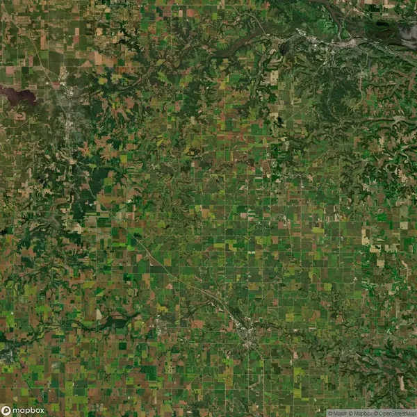 Aerial view of a vast patchwork of green and brown agricultural fields, intersected by roads and streams, with wooded areas visible on the edges.