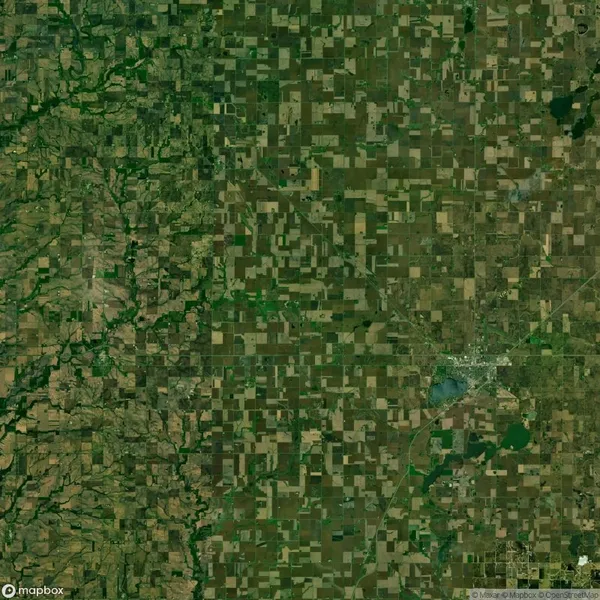 Aerial view of a rural landscape with a patchwork of agricultural fields and a small town. Fields vary in color, arranged in a grid-like pattern. Sparse roads and greenery are visible.