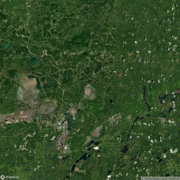 Aerial view of a lush green landscape with scattered clouds, showing a mix of forests, fields, and a few developed areas with visible roads and small water bodies.