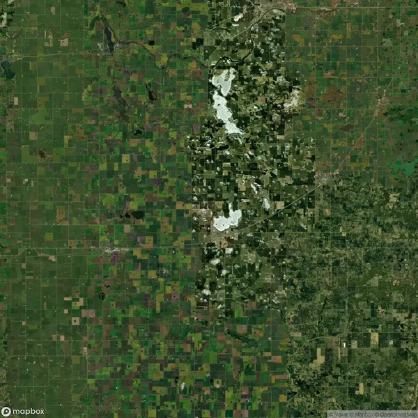 Satellite view of a patchwork landscape with fields and vegetation, featuring a central river or water body, various shades of green, brown, and some urban areas scattered throughout.