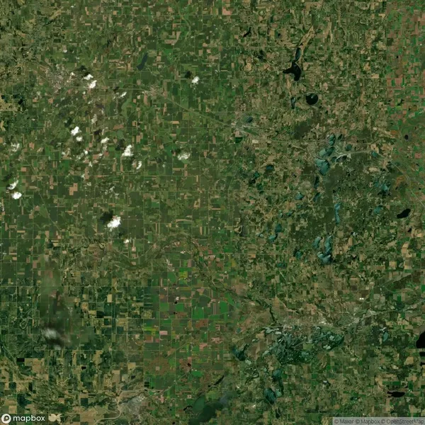 Satellite view of a vast, textured farmland area with various patterns of green fields, scattered clouds, and a few small water bodies.