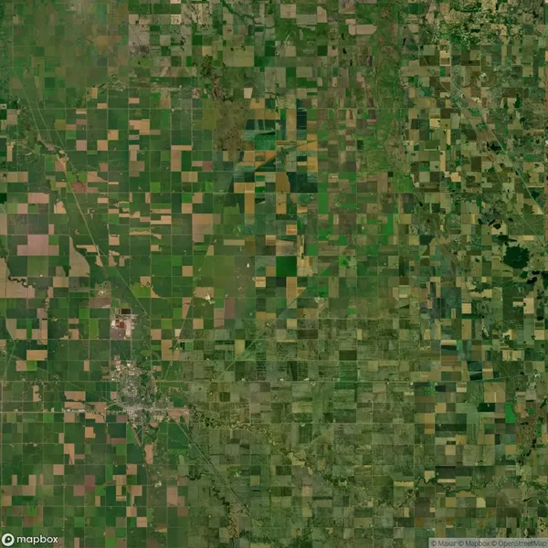 Aerial view of a large, patchwork farmland with various shades of green and brown sections, showing fields, roads, and a small urban area in the lower left corner.