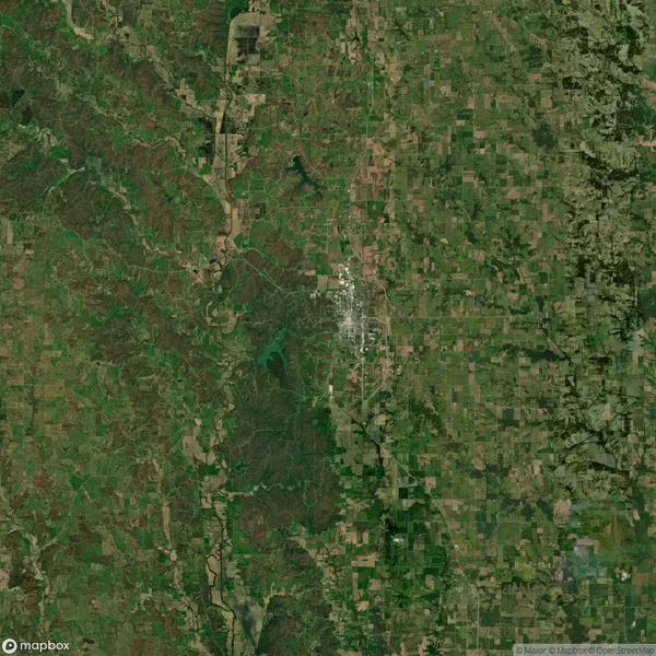 Satellite view of a landscape featuring a dense area of buildings and roads at the center, surrounded by green fields, forests, and patches of agricultural land.