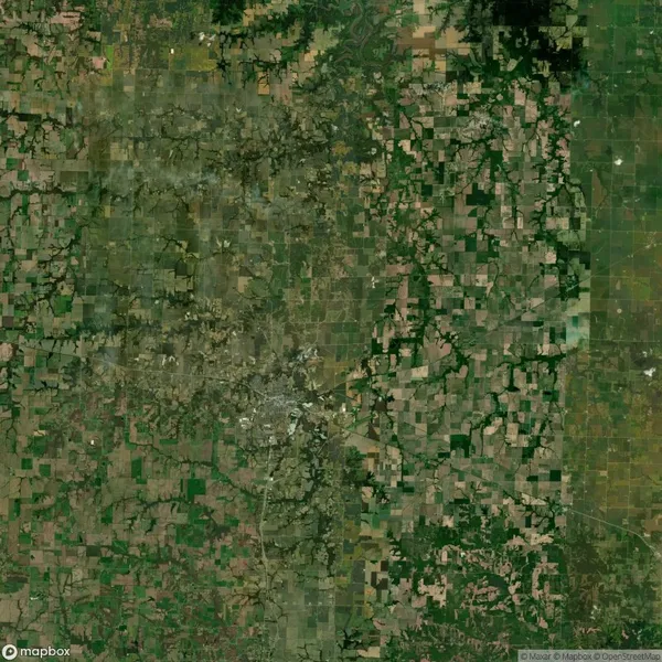 Satellite view of a patchwork landscape with green and brown fields, roads, and a small dense area suggesting urban development in the center.