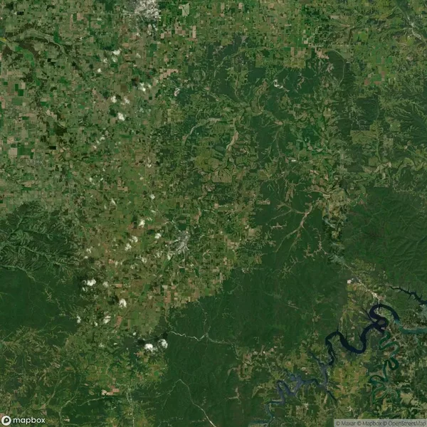 Satellite image showing a landscape with dense green forests, agricultural fields, and a winding river on the bottom right. Small scattered clouds cover part of the area.