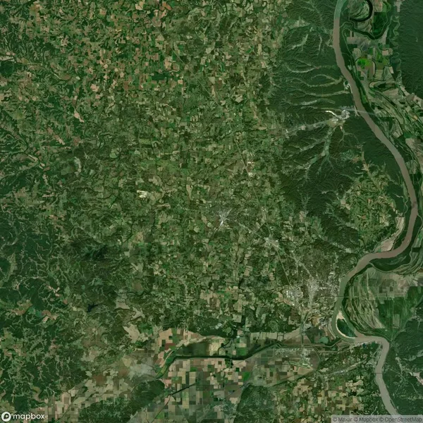 Aerial view of a green landscape with dense forest, agricultural fields, and a winding river on the right. Patches of urban development are visible throughout the terrain.