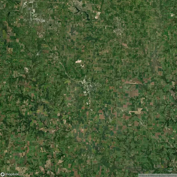Aerial view of a green landscape with patches of farmland and a small urban area at the center. Roads and fields are visible throughout the region.