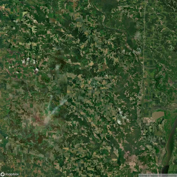 Aerial view of a green, rural landscape with a mix of forests and agricultural fields, crisscrossed by roads. Small cloud formations are visible.
